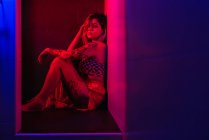Attractive tattooed woman sitting in pink neon light and looking at camera — Stock Photo