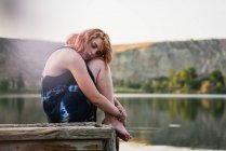 Чуттєва молода жінка сидить на пірсі, обіймаючи коліна в озері в природі — стокове фото
