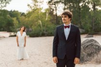 Uomo e donna in eleganti abiti da sposa in piedi separatamente sulla costa sabbiosa con alberi verdi alla luce del sole sullo sfondo — Foto stock