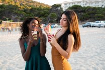 Молодые подруги отдыхают на пляже с напитками в чашках и смеются во время чата на закате — стоковое фото