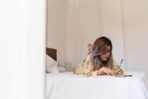 Young woman in silk robe lying on bed and making sketches in notepad in stylish bedroom — Stock Photo