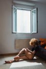 Sensual ethnic woman in bodysuit sitting on floor at home — Stock Photo