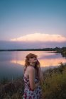 Mujer joven en vestido de verano de pie en la orilla del lago al atardecer y cubriendo los ojos con pelo - foto de stock