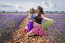 Happy pregnant woman kissing little daughter while sitting on pathway in blooming lavender field — Stock Photo