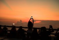 Siluetas de personas tomando fotos con teléfonos inteligentes a la orilla del mar con luces de puesta de sol en Tailandia. - foto de stock