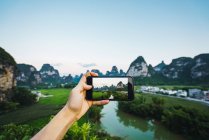 Main humaine prenant des photos avec smartphone de vallée verte avec des montagnes au coucher du soleil, Guangxi, Chine — Photo de stock
