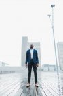 Homem de negócios elegante em pé no pavimento na chuva contra edifícios modernos da cidade — Fotografia de Stock