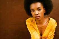 Femme afro-américaine en robe jaune vif regardant la caméra sur fond brun — Photo de stock