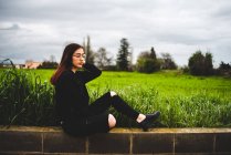 Портрет стильной молодой женщины, сидящей на кирпичной стене в загородной местности в пасмурную погоду — стоковое фото