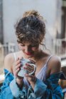 Young curly girl in white underwear and denim jacket standing on balcony with coffee cup looking dreamily away — Stock Photo
