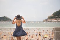 Woman holding hat and enjoying view of crowded beach and calm sea while standing near fence — Stock Photo