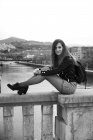 Side view of young attractive woman in leather jacket and short skirt with tattoo on leg and backpack sitting on stone parapet of bridge in Bilbao and looking at camera on background of river — Stock Photo
