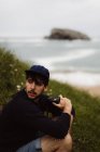 Young man sitting on grass on coast and looking away while holding camera in hand and sea on background in Cantabria, Spain — Stock Photo