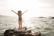 Frau in Badebekleidung steht an sonnigen Tagen mit erhobenen Händen auf Felsbrocken im Meer — Stockfoto