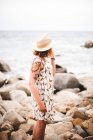 Вид ззаду татуйованої жінки в капелюсі, що стоїть на каменях з розбитими руками і дивиться на океан — стокове фото