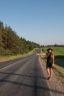 Jovem mulher vestindo vestido e chapéu segurando o polegar para cima enquanto carona na beira da estrada do campo — Fotografia de Stock