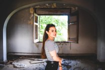 Attrayant jeune femme en tenue décontractée regardant la caméra tout en se tenant dans le couloir de vieux bâtiment abandonné. — Photo de stock