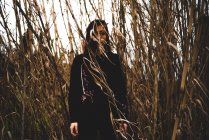 Mysterious girl wearing black outfit standing in tall dried grass and looking at camera — Stock Photo