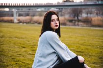Morena séria em roupa casual e com maquiagem escura sentada na grama verde olhando para a câmera. — Fotografia de Stock