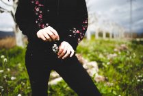 Fille en tenue noire tenant petite branche de fleurs tout en se tenant debout dans le champ en fleurs — Photo de stock