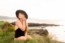 Junge Frau in schwarzem Badeanzug und Hut am Meer sitzend — Stockfoto