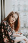 Portrait of Young redheaded woman standing at window — Stock Photo