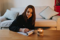 Молода жінка пише в блокноті за столом зі свічкою вдома — стокове фото
