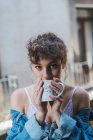 Young curly girl in white underwear and denim jacket standing on balcony with coffee cup looking dreamily away — Stock Photo