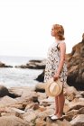 Back view of tattooed woman in hat standing on stones with hands apart and looking at the ocean — Stock Photo