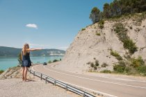 Mujer joven autostop en la carretera de carretera de carretera estrecha en el mar - foto de stock