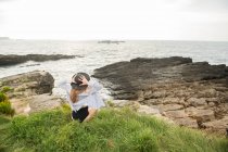 Frau mit Hut sitzt auf Gras an der Küste und genießt den Blick aufs Meer — Stockfoto