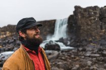 Bello uomo barbuto guardando lontano mentre in piedi sullo sfondo di una bella cascata durante il viaggio attraverso l'Islanda. — Foto stock