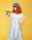 Изолированный вид рыжеволосой модели в синем платье и искусственных цветов на голове, поднимая руки, держа и глядя на хризантему на желтом фоне — стоковое фото