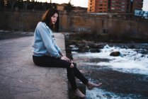 Side view of casual brunette in hoodie sitting barefoot on concrete waterfront with waves below and looking away. — Stock Photo
