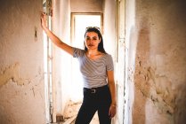 Attractive young woman in casual outfit looking at camera while standing in corridor of old abandoned building. — Stock Photo