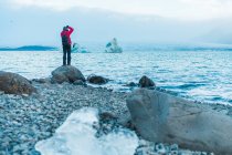 Задний вид мужчины в теплой одежде и рюкзаке, фотографирующего живописное озеро Скафтафель, ледник и ватнайокулл — стоковое фото