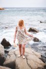 Visão traseira da mulher tatuada de chapéu em pé sobre pedras com as mãos afastadas e olhando para o oceano — Fotografia de Stock
