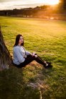 Vista lateral da morena casual sentada sob a árvore no gramado contra a cidade no pôr do sol e no céu sombrio. — Fotografia de Stock