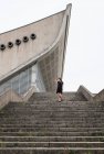 Знизу знімок жінки в чорному платті, що йде сірими бетонними сходами з сучасною будівлею на фоні — стокове фото