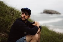Giovane seduto sull'erba sulla costa e distogliendo lo sguardo mentre tiene la fotocamera in mano e in mare sullo sfondo in Cantabria, Spagna — Foto stock