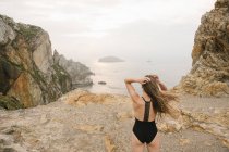 Woman in black swimsuit standing on rocky shore and looking at view — Stock Photo