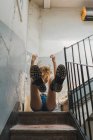 De baixo de menina encaracolado em shorts e botas sentado em passos com cachos voando e rindo em movimento — Fotografia de Stock