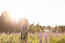 Mulher anônima em chapéu e vestido de pé com um monte de flores em? florescendo campo em brilhante de volta iluminado — Fotografia de Stock