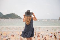 Резервного зору молода жінка тримає капелюх, насолоджуючись видом на переповненому пляжі та спокійне море, поки стоїть біля огорожі — стокове фото