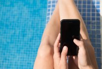 Mains féminines à l'aide d'un smartphone avec écran vide tout en étant assis près de la piscine avec de l'eau propre — Photo de stock