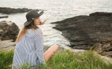 Молода жінка в капелюсі і сорочку, сидячи на траві на узбережжі і насолоджуватися видом на море — стокове фото