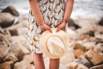 Vista posterior de la mujer de la cosecha sosteniendo sombrero y de pie en la costa rocosa en el océano - foto de stock