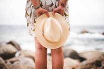 Задний вид женщины урожая, держащей шляпу и стоящей на скалистом побережье у океана — стоковое фото