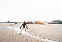 Donna scalza che tiene cappello e calzature mentre salta sopra il flusso sulla spiaggia sabbiosa — Foto stock