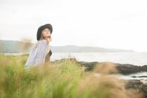 Mujer joven reflexiva en sombrero y camisa sentado en la hierba en la costa con los ojos cerrados - foto de stock